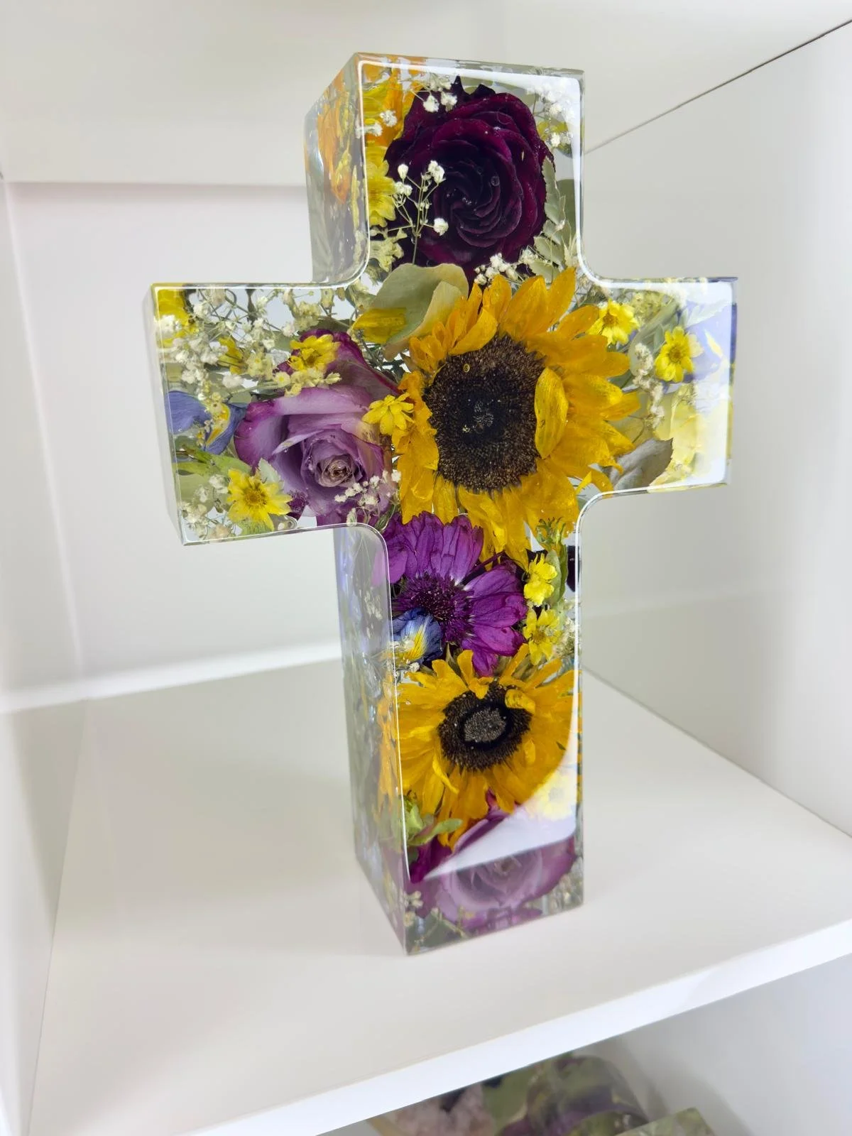 A resin cross-shaped vase filled with colorful flowers including sunflowers, purple roses, and yellow and purple daisies.