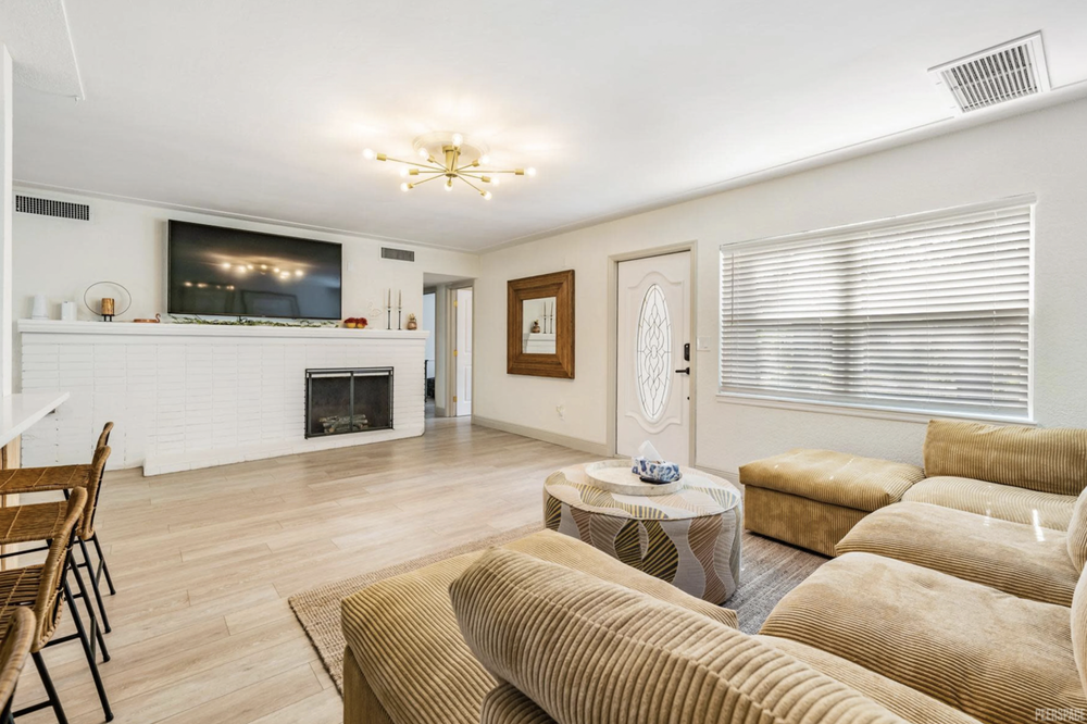 Living room with beige sectional sofa, round coffee table, large window with blinds, white door, and a wall-mounted TV above a white fireplace