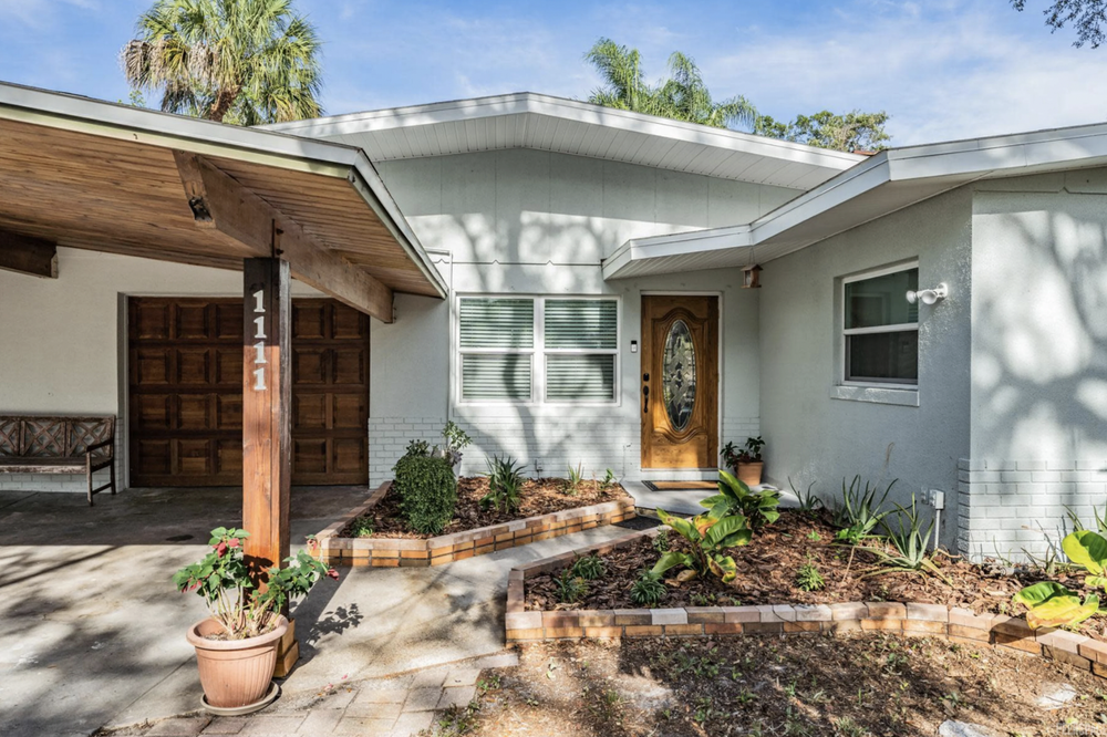 Front view of a single-story house with a wooden front door, a garage, and a small garden with plants and shrubs.