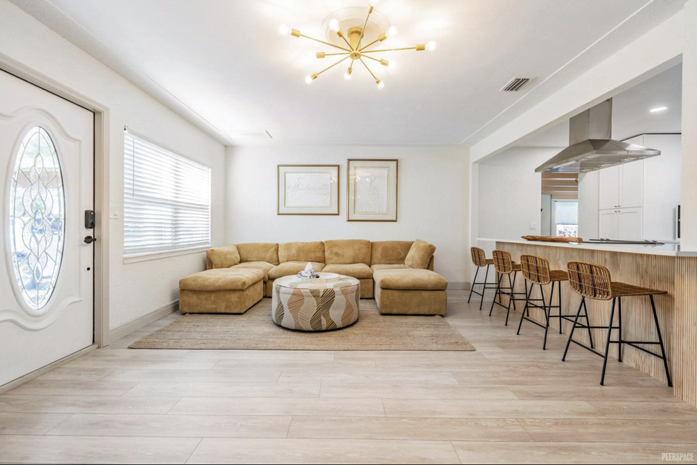 Living room with a beige sectional sofa, a round patterned ottoman, a beige area rug, and wall art. Adjacent kitchen area with a bar counter and five wicker bar stools. Bright lighting and wood flooring.