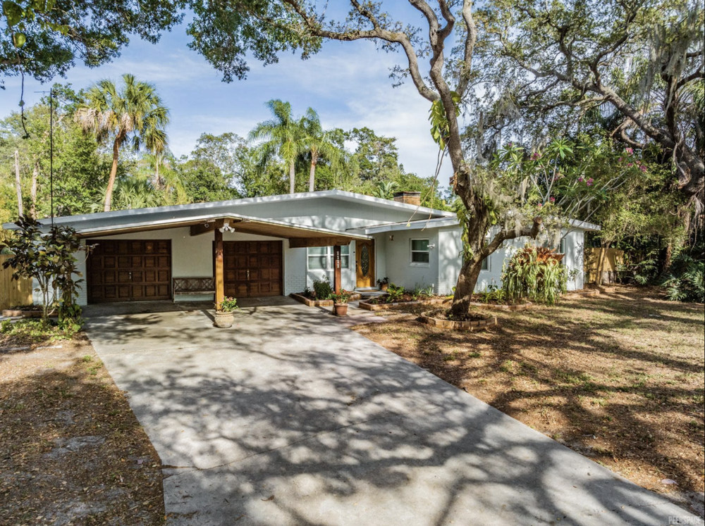 A single-story house with a concrete driveway, surrounded by trees, with tall palm trees in the background and potted plants near the entrance.