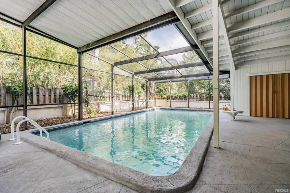Indoor swimming pool with screened enclosure, concrete deck, and wooden cabinet, surrounded by trees and fence.