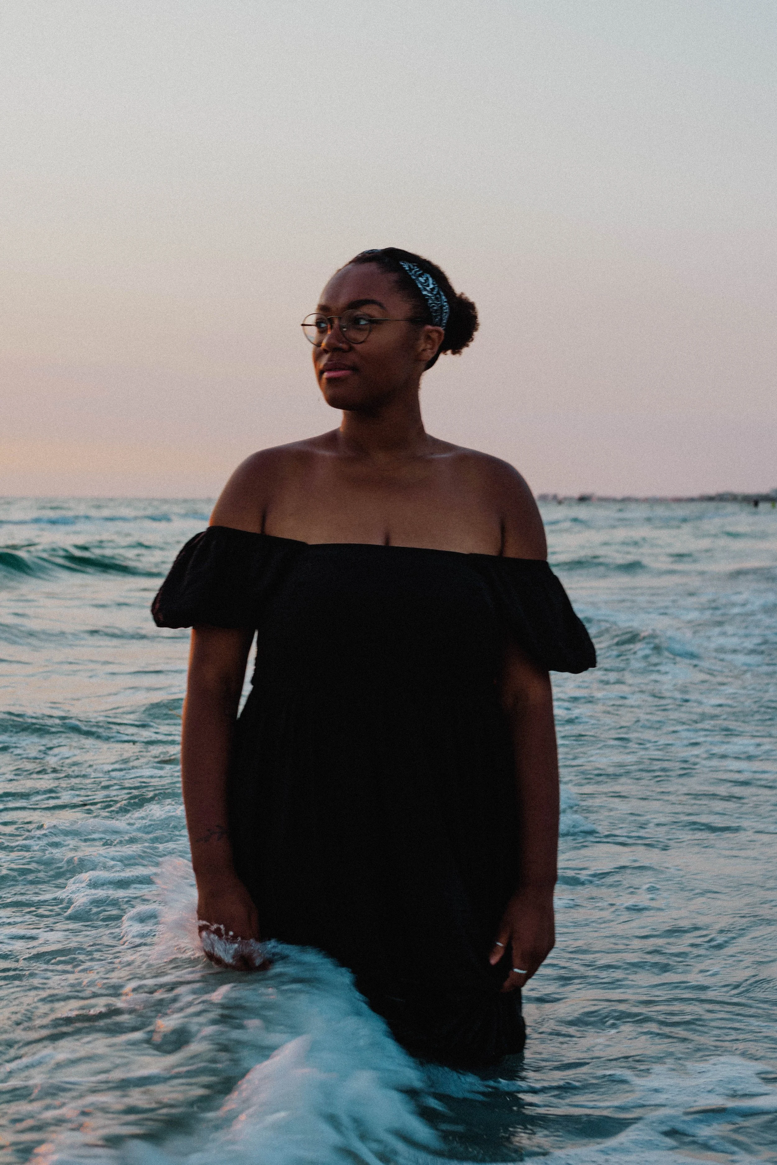 A woman standing in the ocean during sunset, wearing a black off-the-shoulder dress, glasses, and a blue headband.