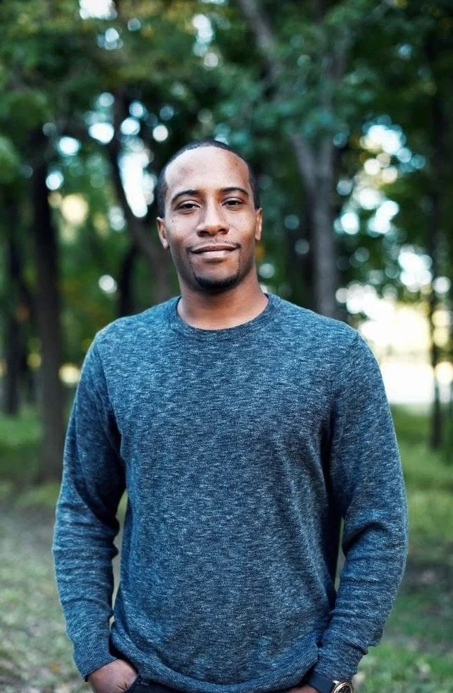 A man standing outdoors in front of trees, wearing a dark blue sweater, with his hands in his pockets, looking at the camera and smiling slightly.