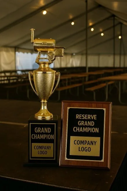 A large golden trophy and an award plaque on a table inside a tented event space. The trophy has a gold laser gun on top, and both awards have placeholder text indicating "Grand Champion" and "Reserve Grand Champion" with space for a company logo.