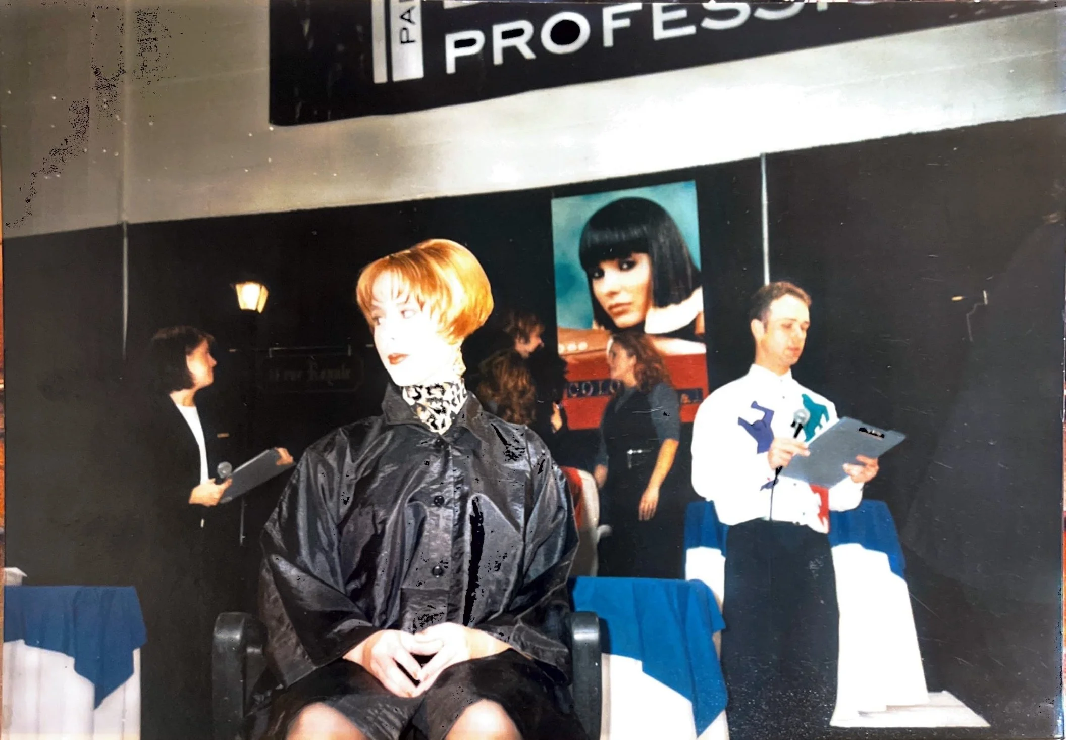 A woman with short red hair wearing a black jacket and leopard print scarf seated in the foreground. In the background, a man in a white suit with colorful patches on his sleeves holding a paper and microphone, a woman with dark hair holding a microphone and clipboard, and several other people are visible. There are large posters on the wall, including one of a woman with a black bob haircut and a poster with the word 'PROFESS' partially visible.