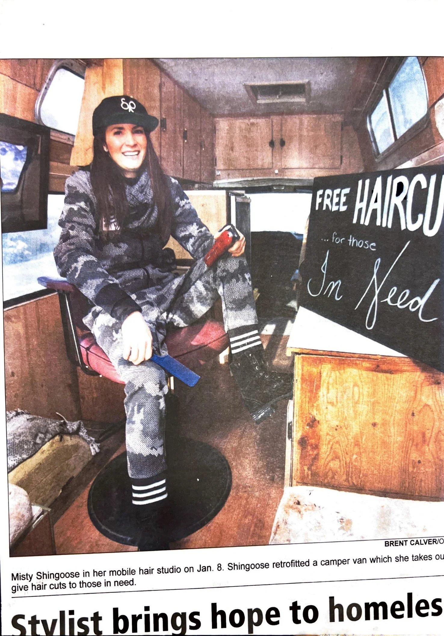 A woman sitting inside a vintage camper van, holding a haircutting tool, with a sign that reads 'Free Haircuts for those in Need.' She wears camouflage-patterned clothing and a black cap, smiling at the camera.