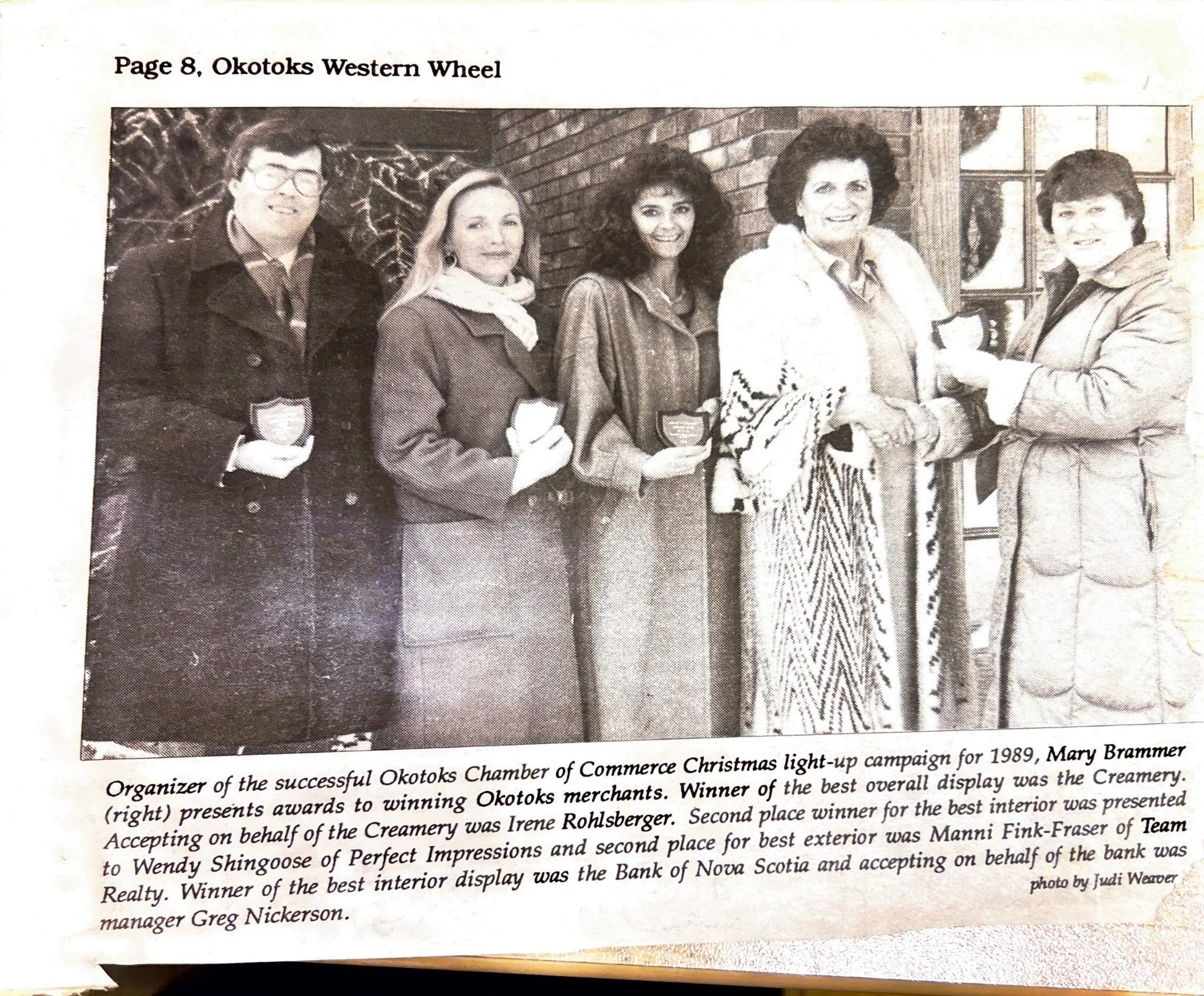 Black and white photo of five women and one man standing outside by a brick building, with three women holding awards. A woman in the middle is shaking hands with another woman. The photo is part of a newspaper page titled "Page 8, Okotoks Western Wheel" and includes a caption describing the event.