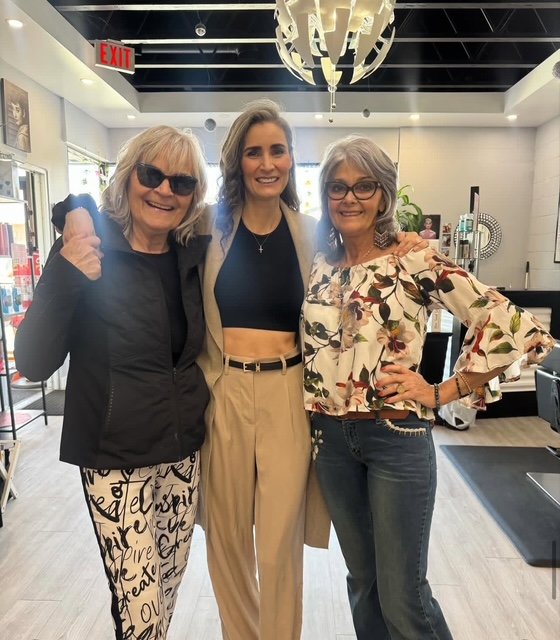Three women smiling and posing together indoors, one with sunglasses and two wearing glasses, in a modern space with a chandelier and various decor. Beauty, happiness, professionals, small business owners, support, hair stylists, perms, hair colour,