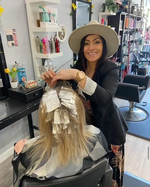 A woman in a large hat and blazer is styling a client's long, blonde hair with foils in a salon.
