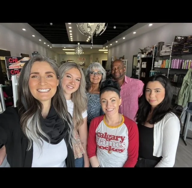 Group of six people smiling and posing for a photo inside a well-lit store or library with shelves of books and products in the background. Barbershop, cuts, colors, perms, extensions, professionals, Okotoks, Elizabeth Street, Shopping, Spa