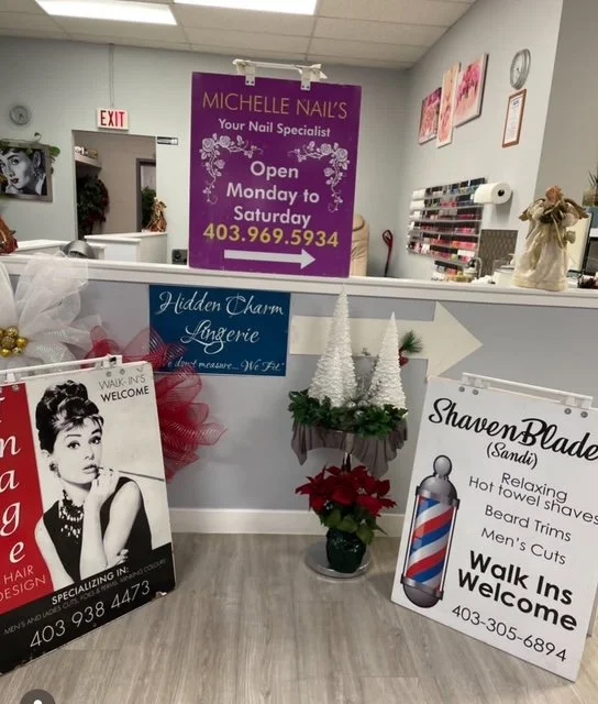 Inside a nail salon with a reception desk, decorative holiday trees on a plant, and various signage advertising services like waxing, lingerie, and shaving blades, along with phone numbers.