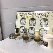 Display of vintage shaving brushes on a stand labeled "Famous Shaving Styles" with six brushes and a container of shaving cream or soap.