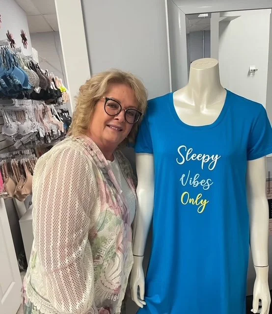 A woman in glasses and a patterned light-colored shirt smiling next to a mannequin wearing a blue t-shirt with the words "Sleepy Vibes Only" written on it.
