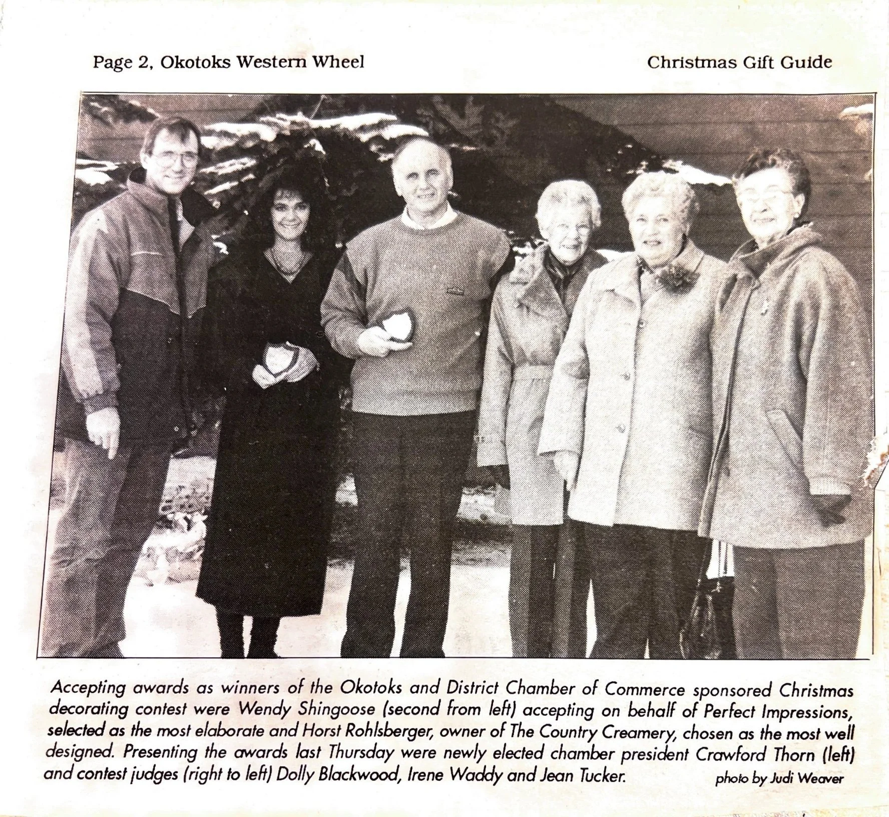 Black and white photograph of seven people standing outdoors with mountains in the background. They are dressed in winter clothing, and some are holding awards.