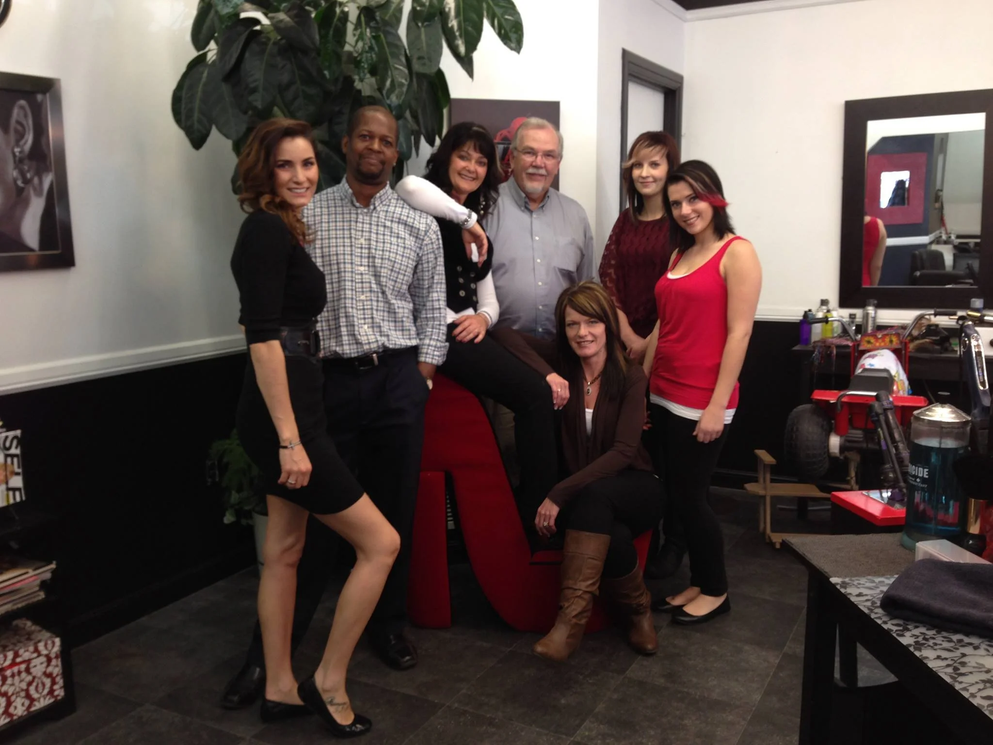 Group of eight people posing together in an indoor setting, standing around a red chair, with a large green plant and framed pictures on the wall in the background.