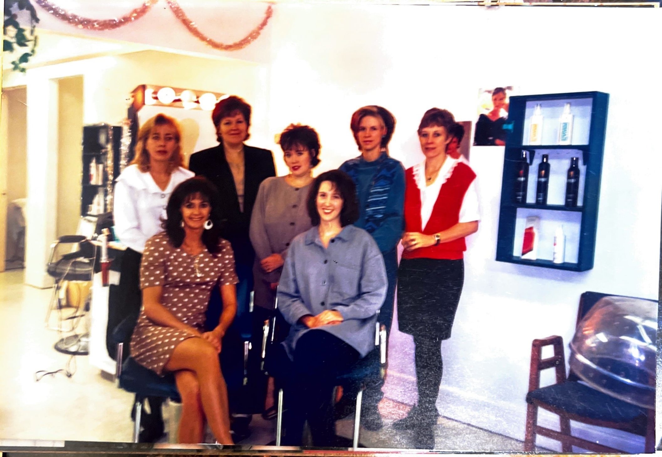 Group of seven women posing together in an indoor setting, some seated and others standing, with salon products on display and salon equipment visible in the background.
