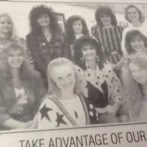 Black and white photo of ten young women posing for a group picture, some wearing dresses and others in casual clothing, with a partial banner reading 'TAKE ADVANTAGE OF OUR' visible in front.