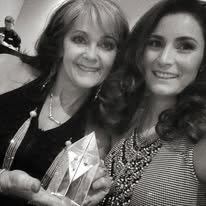 Two women smiling, one older and one younger, at a social event with awards or trophies in front of them.