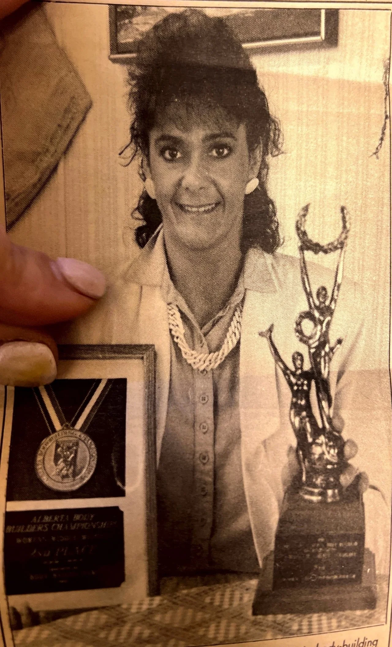 Black and white photo of a woman with short, curly hair, wearing a light-colored blazer, a patterned scarf, and earrings. She is smiling and standing next to a trophy and a framed award, with a small sculpture of dancers in front of her.