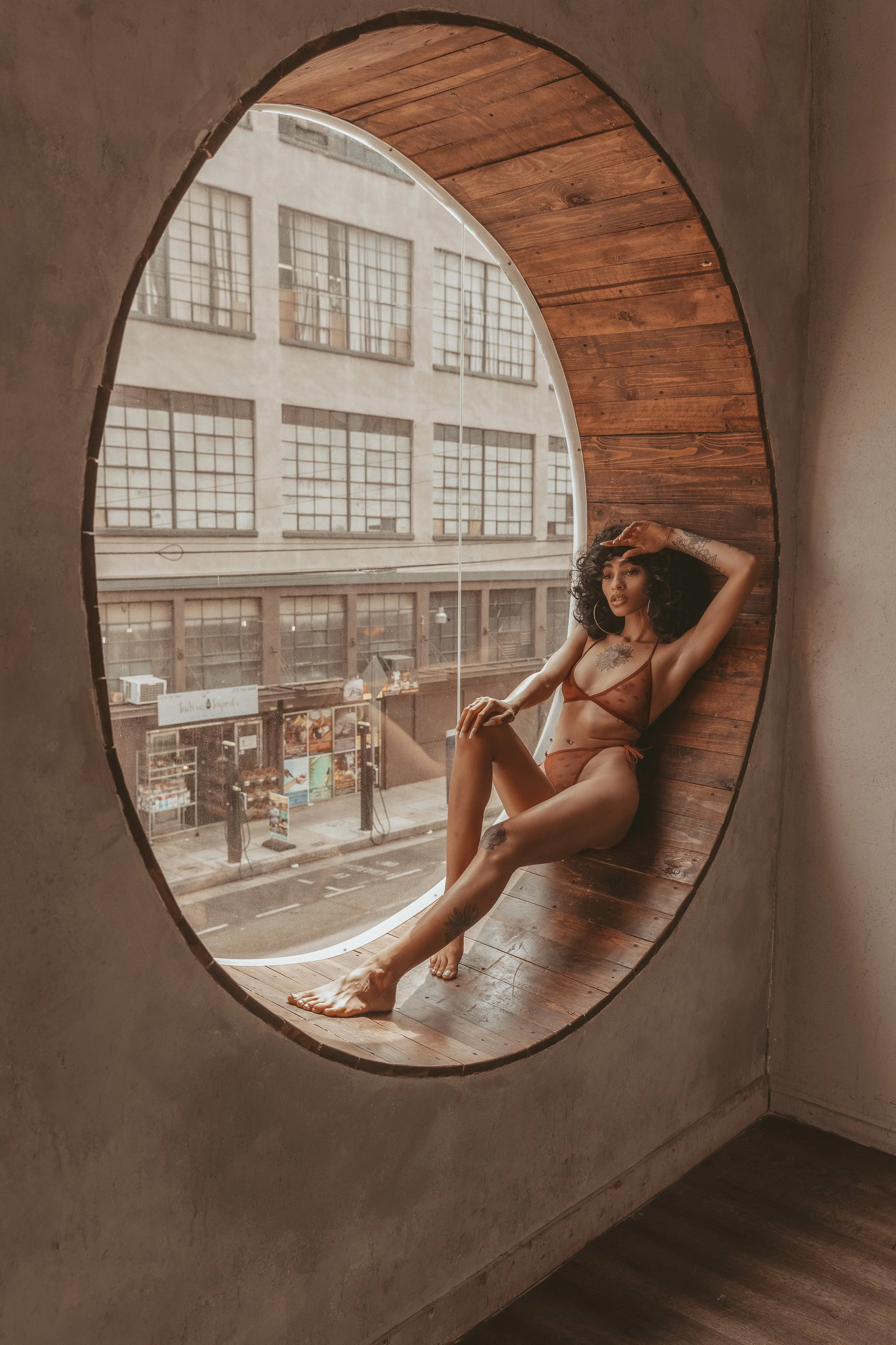 A woman with curly hair and tattoos sitting inside a circular window nook, with an urban city street outside.