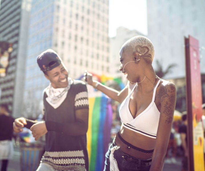 Two women dancing and smiling outdoors at a city event, with tall buildings in the background and rainbow flags.