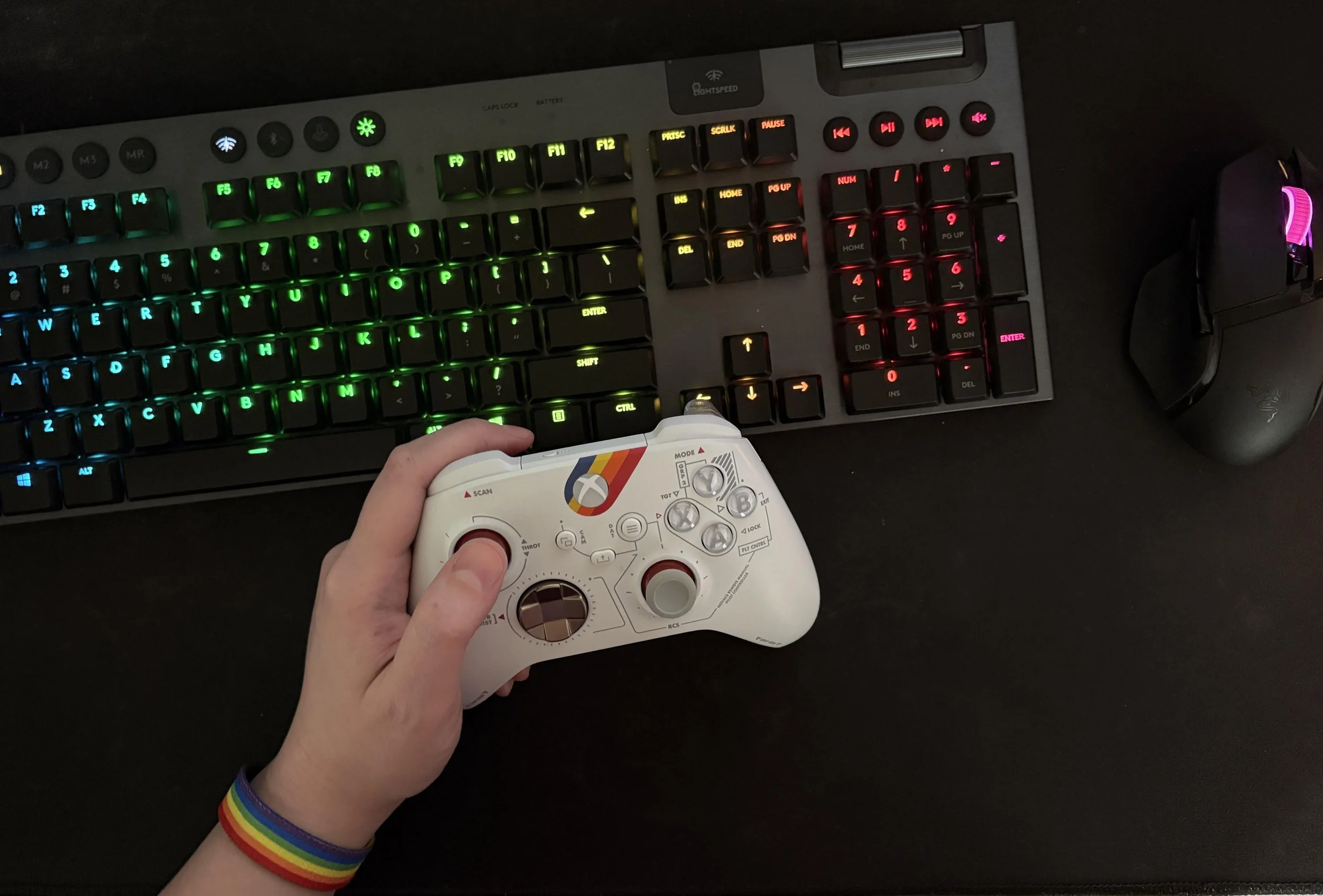 A computer keyboard with colorful backlit keys, a gaming mouse, and a white gaming controller being held in a person's left hand, which is also wearing a rainbow wristband.