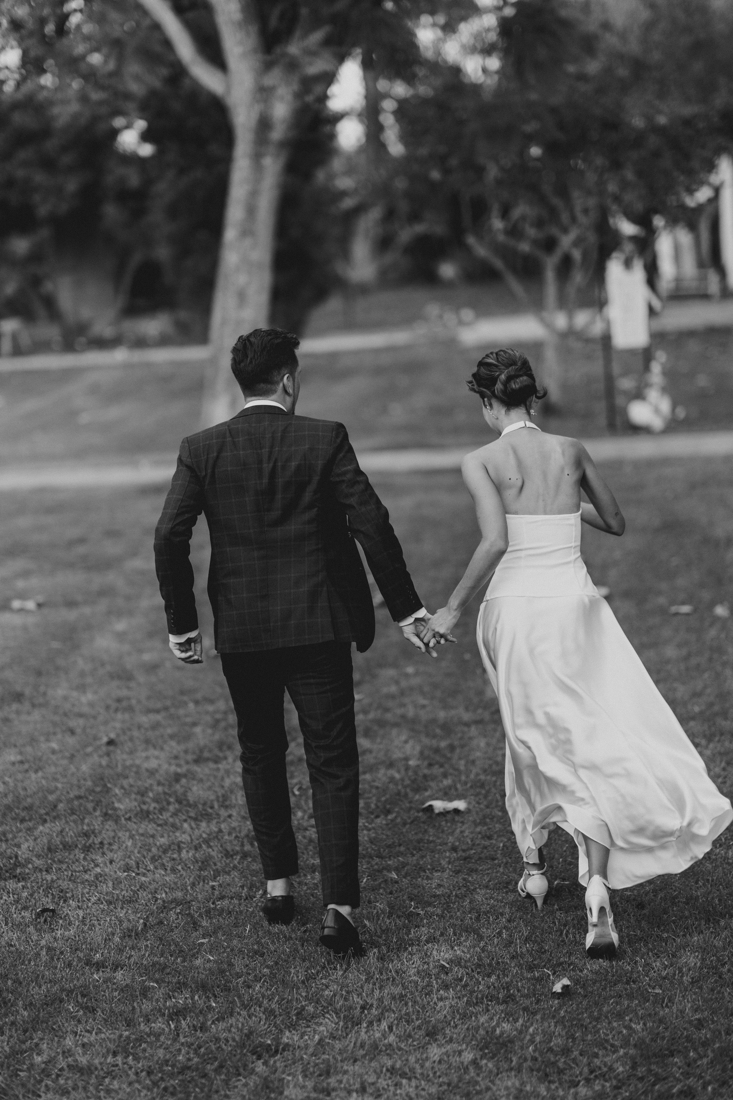 A black and white photo of a couple walking hand-in-hand through a park, dressed in formal attire. The man is wearing a dark, checkered suit and the woman is in a strapless, flowing white gown.