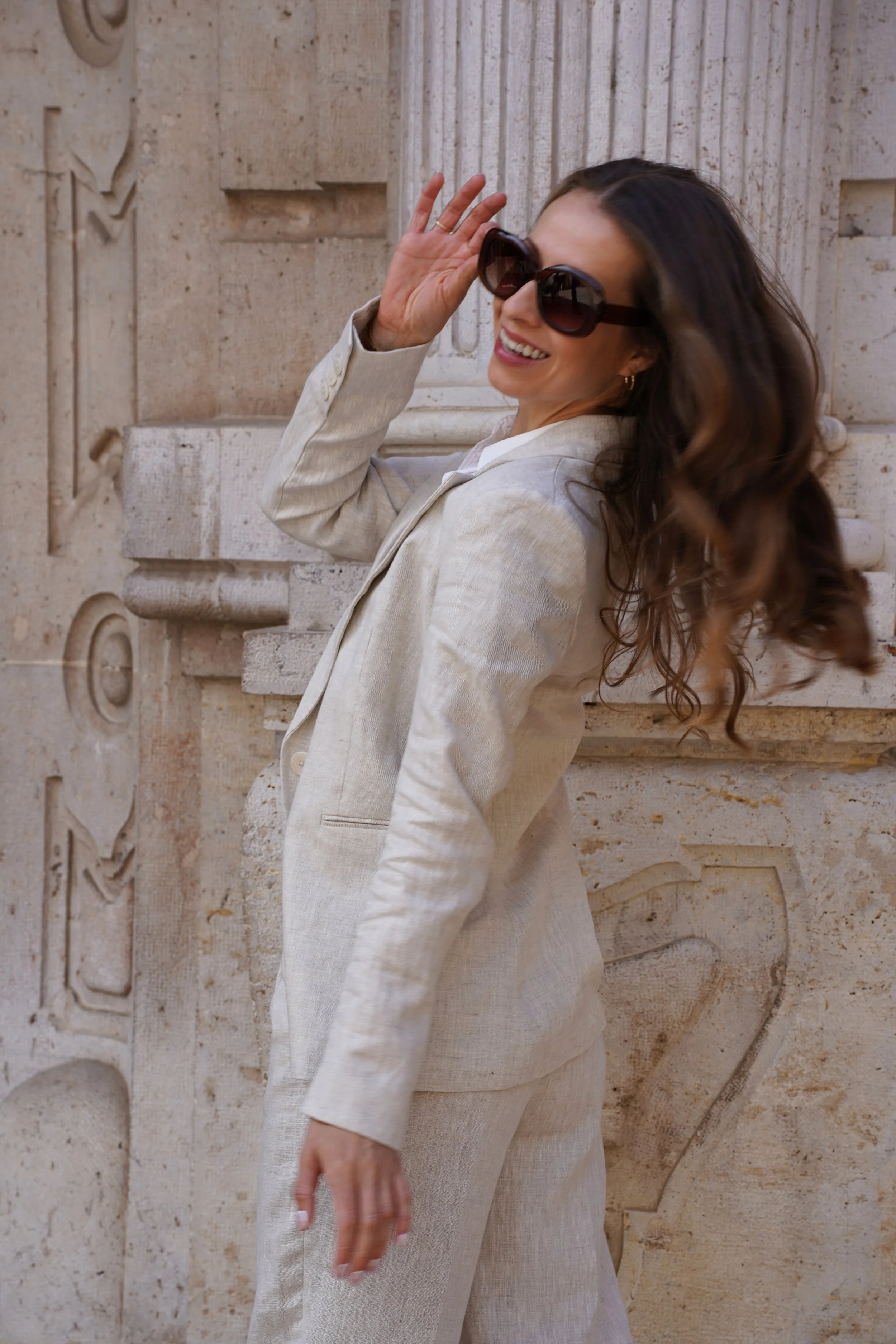 A woman smiling and wearing large sunglasses, a light-colored suit, standing outside near a stone wall.