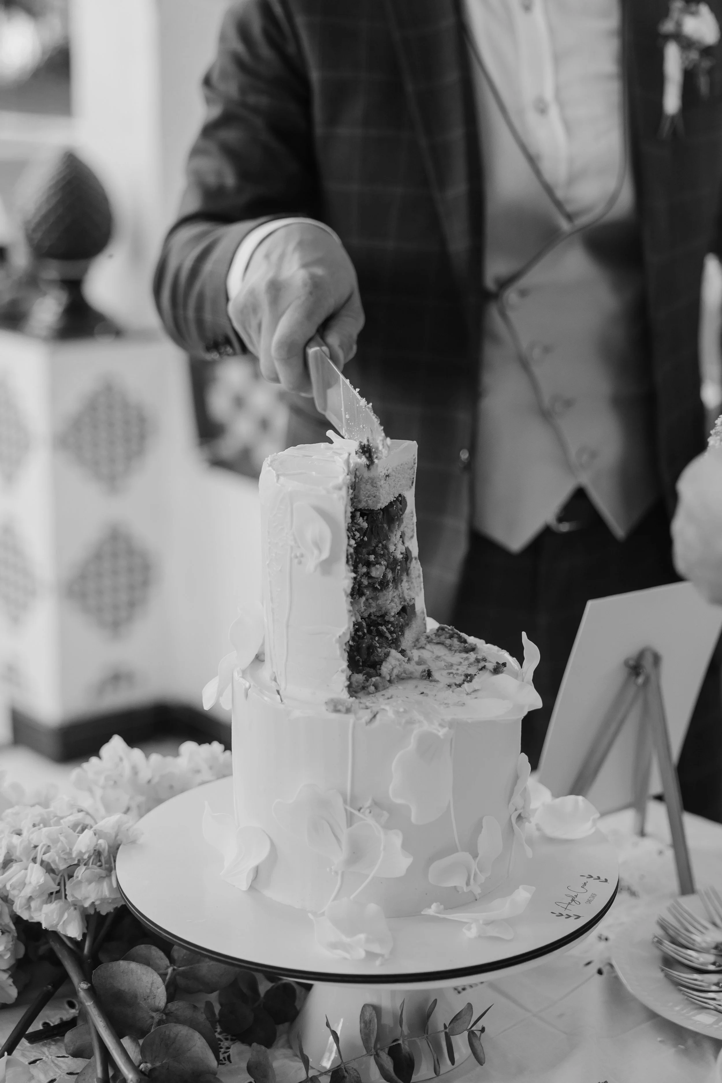 Black and white photo of a person in a suit cutting a slice from a decorated wedding or celebration cake with flowers and leaves around it.