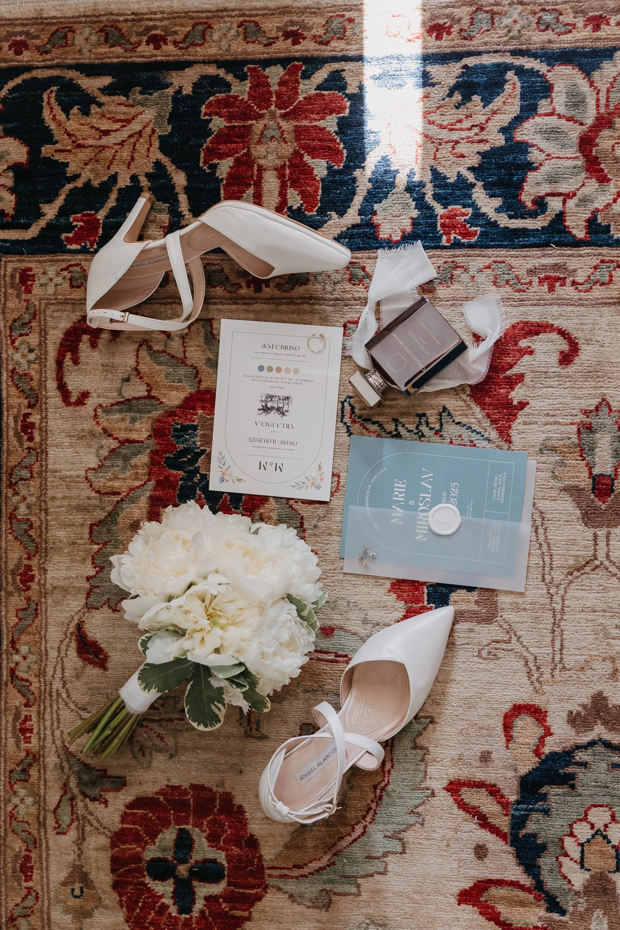 White wedding shoes, a bouquet of white flowers, wedding programs, and other wedding accessories arranged on a patterned rug.