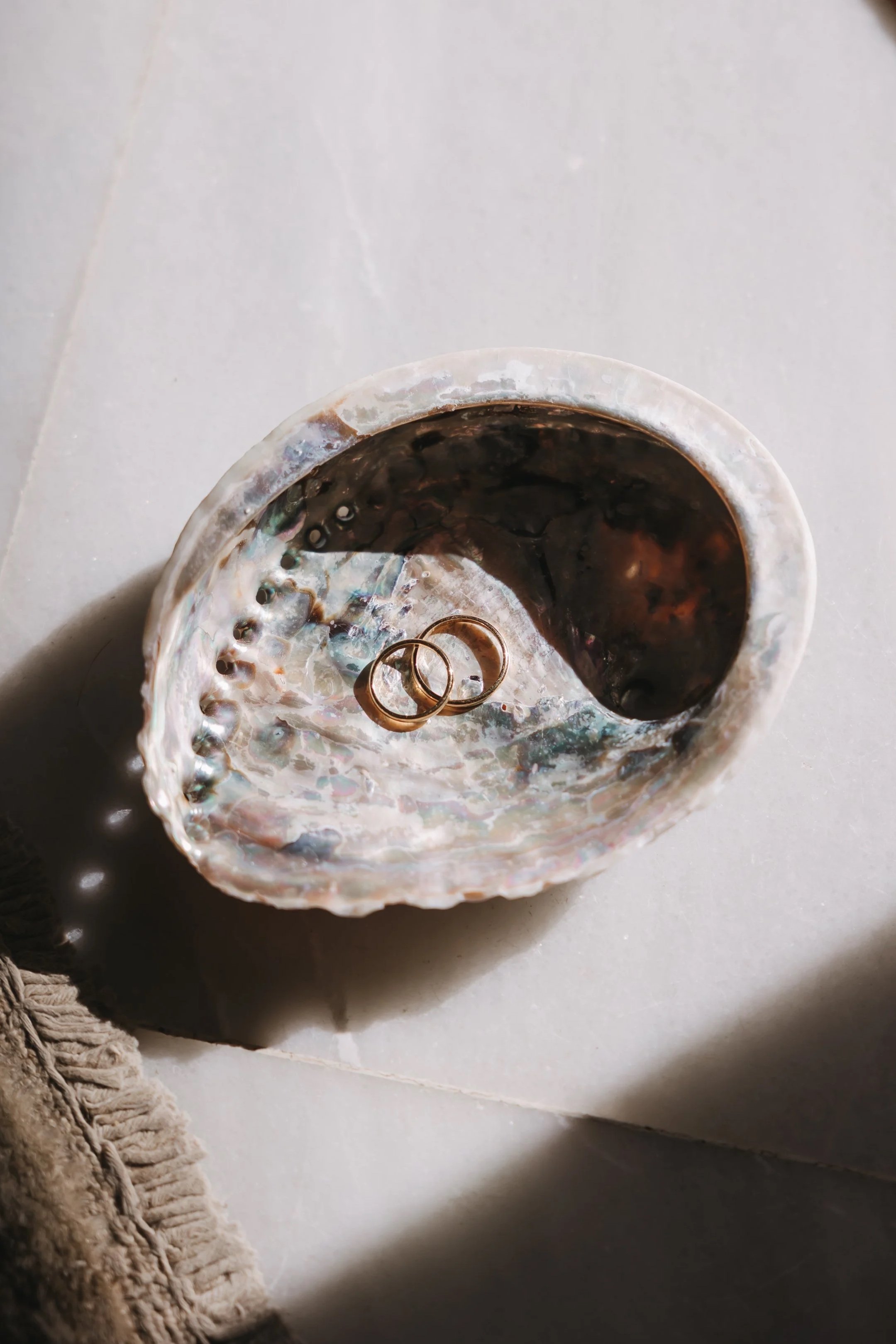 A seashell with two gold rings inside, placed on a tiled surface with shadows and a piece of beige fabric in the corner.