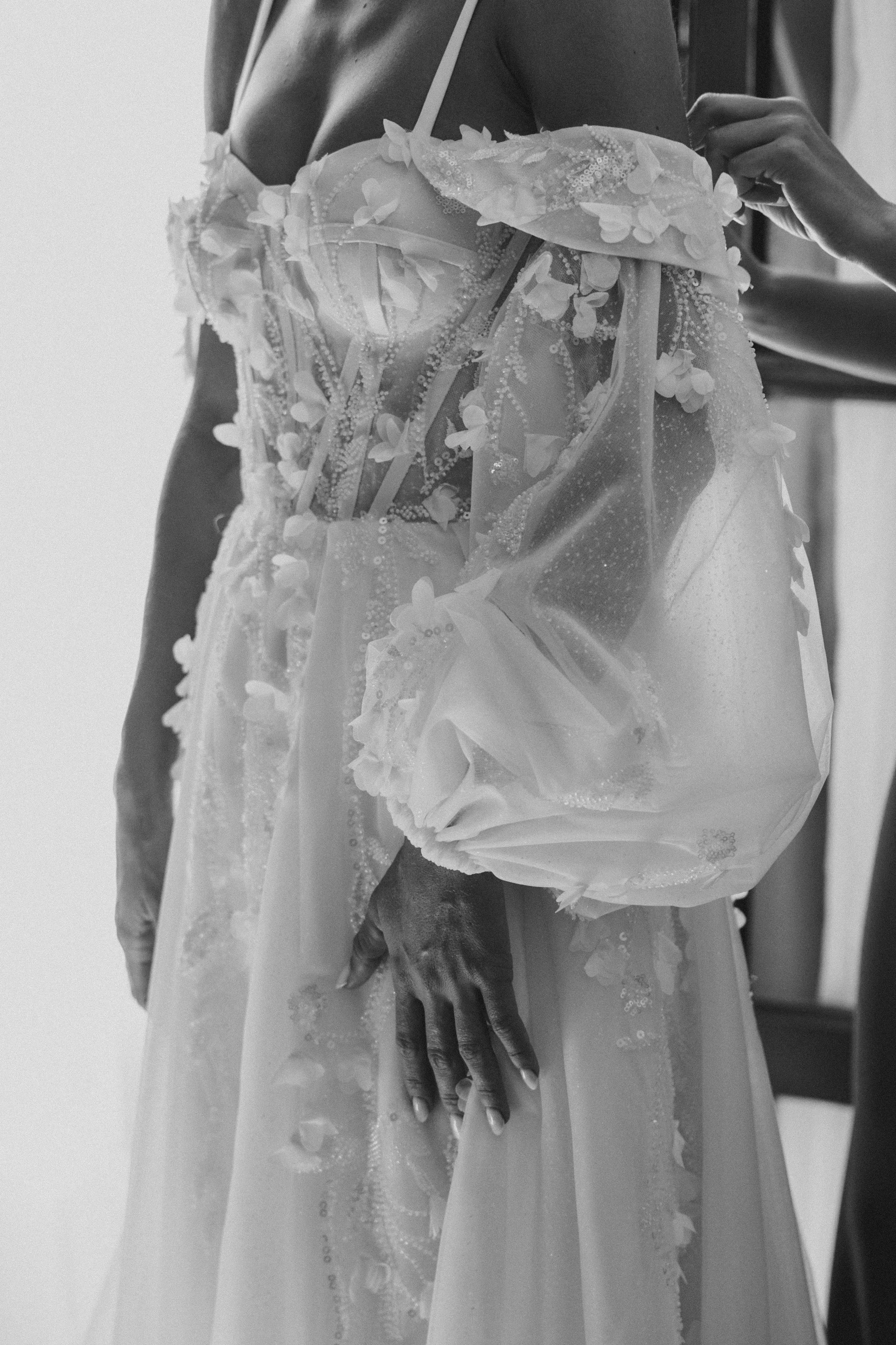 A woman wearing a wedding dress with floral details and transparent fabric, adjusting the off-shoulder sleeve.