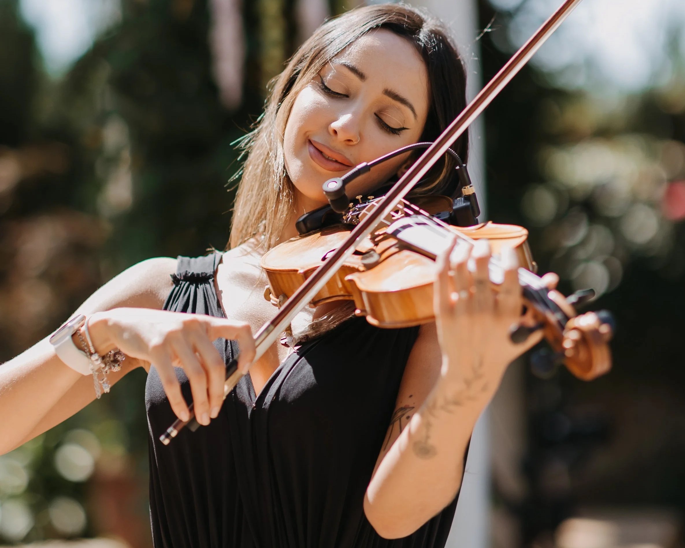 A young woman playing a violin outdoors, wearing a black dress, with a joyful expression and a focused posture.