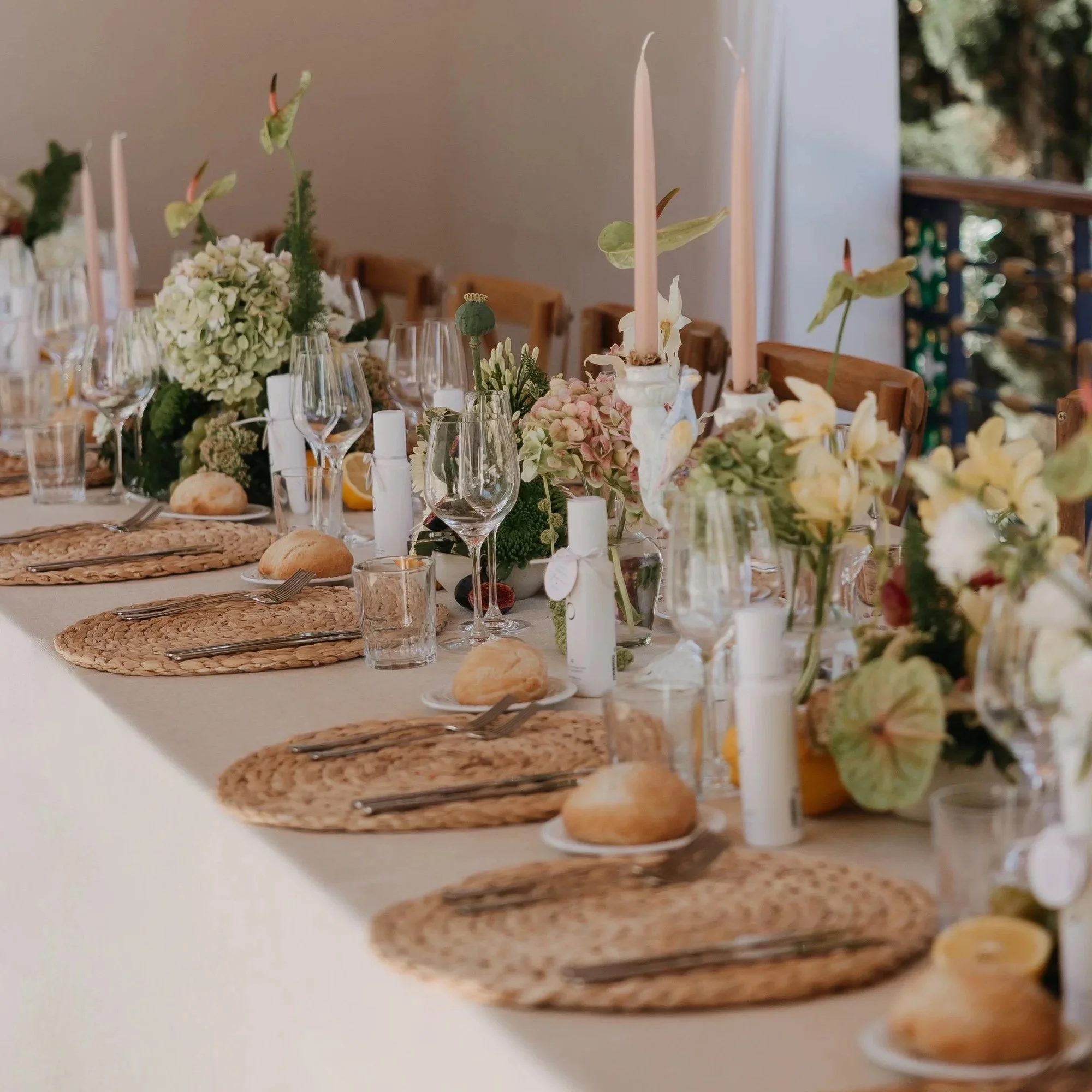 A decorated dining table set with plates, cutlery, wine glasses, candles, bread rolls, and floral centerpieces with pink flowers and green foliage. The setting suggests a formal or special occasion.