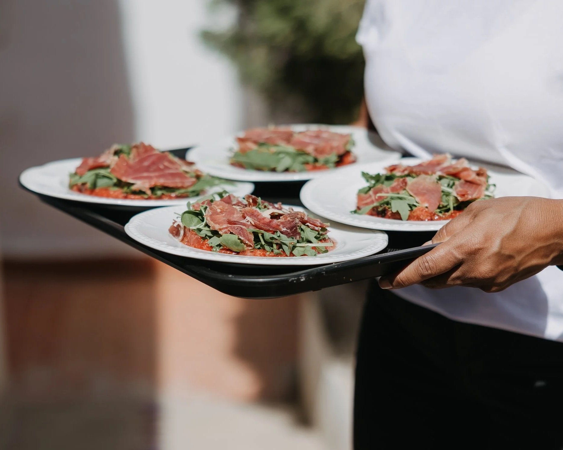 A person holding a black tray with four plates of food, each topped with arugula and cured ham slices, outdoors.