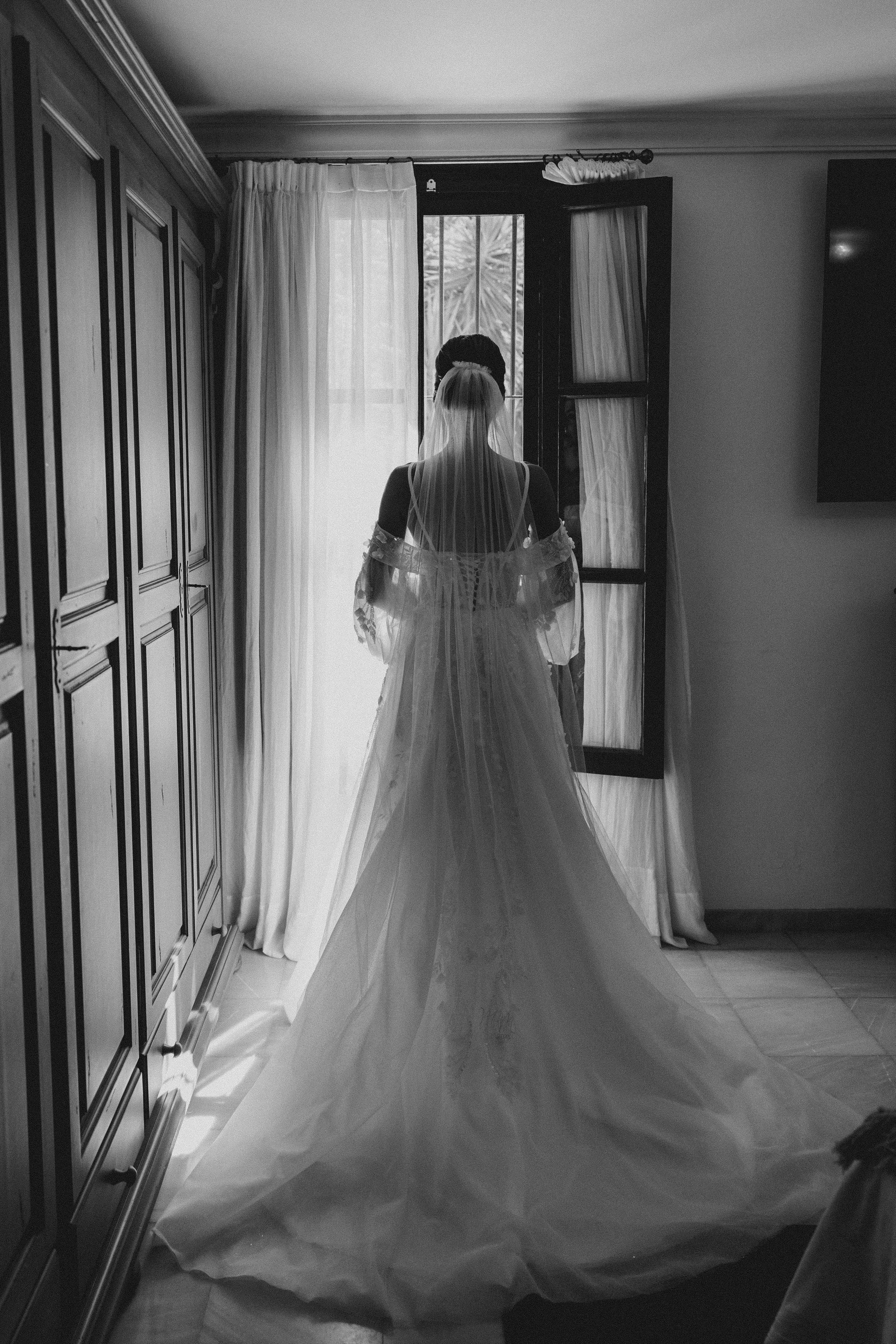 Bride in wedding dress with veil standing by window in a room.