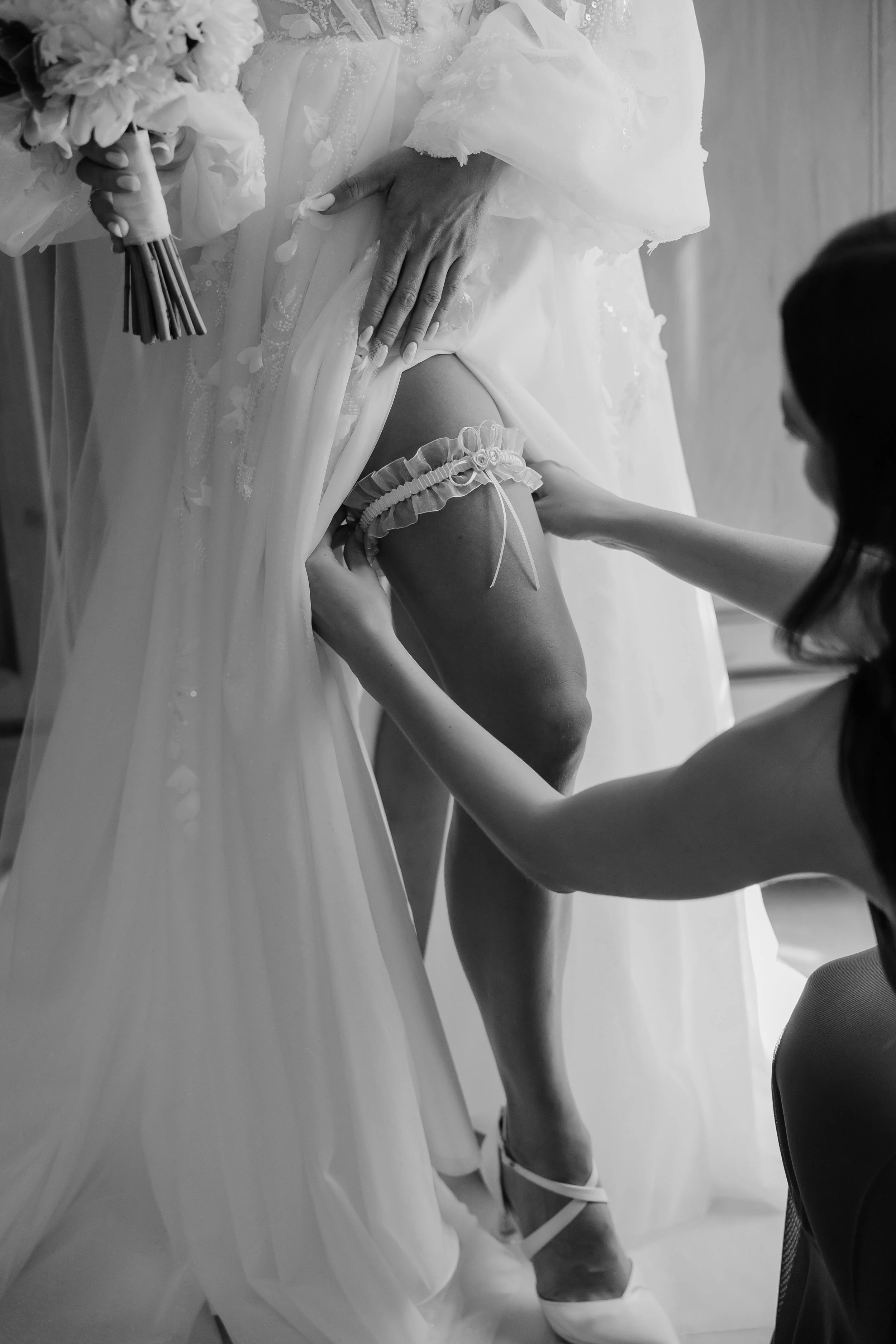 A bride getting her stocking adjusted while another woman helps, with a bouquet in her hand. The scene is in black and white.