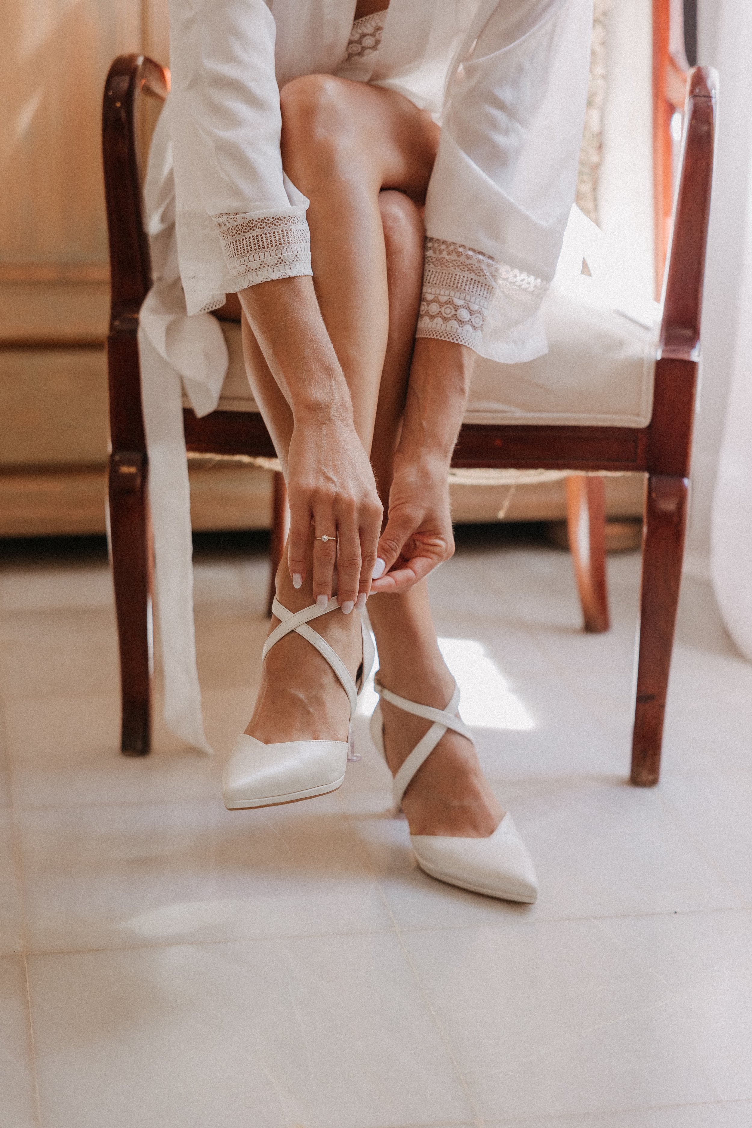 A woman is sitting on a wooden chair, adjusting her white high heels while wearing a white robe with lace cuffs.