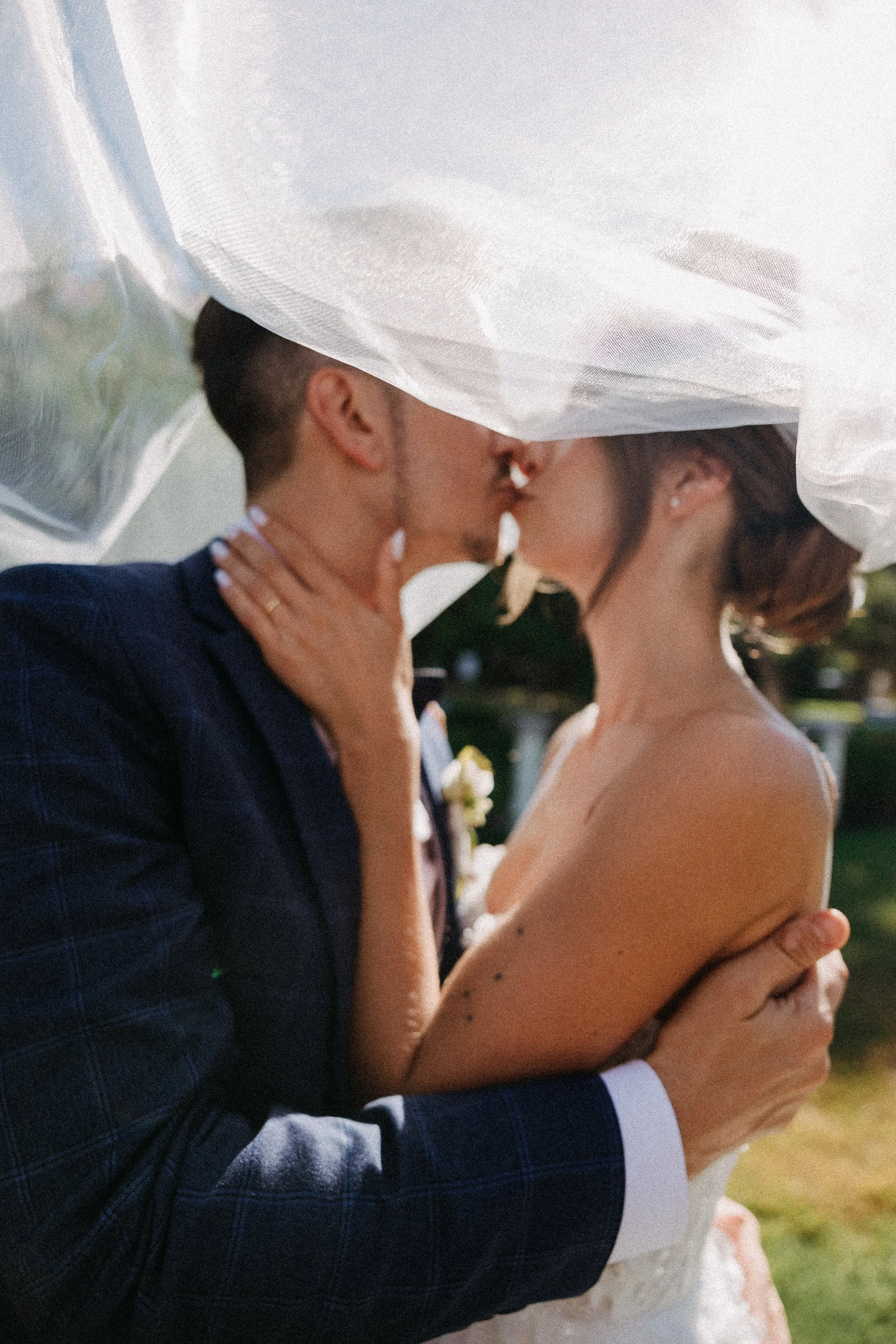 A bride and groom sharing a kiss under a veil during their wedding ceremony in an outdoor setting.
