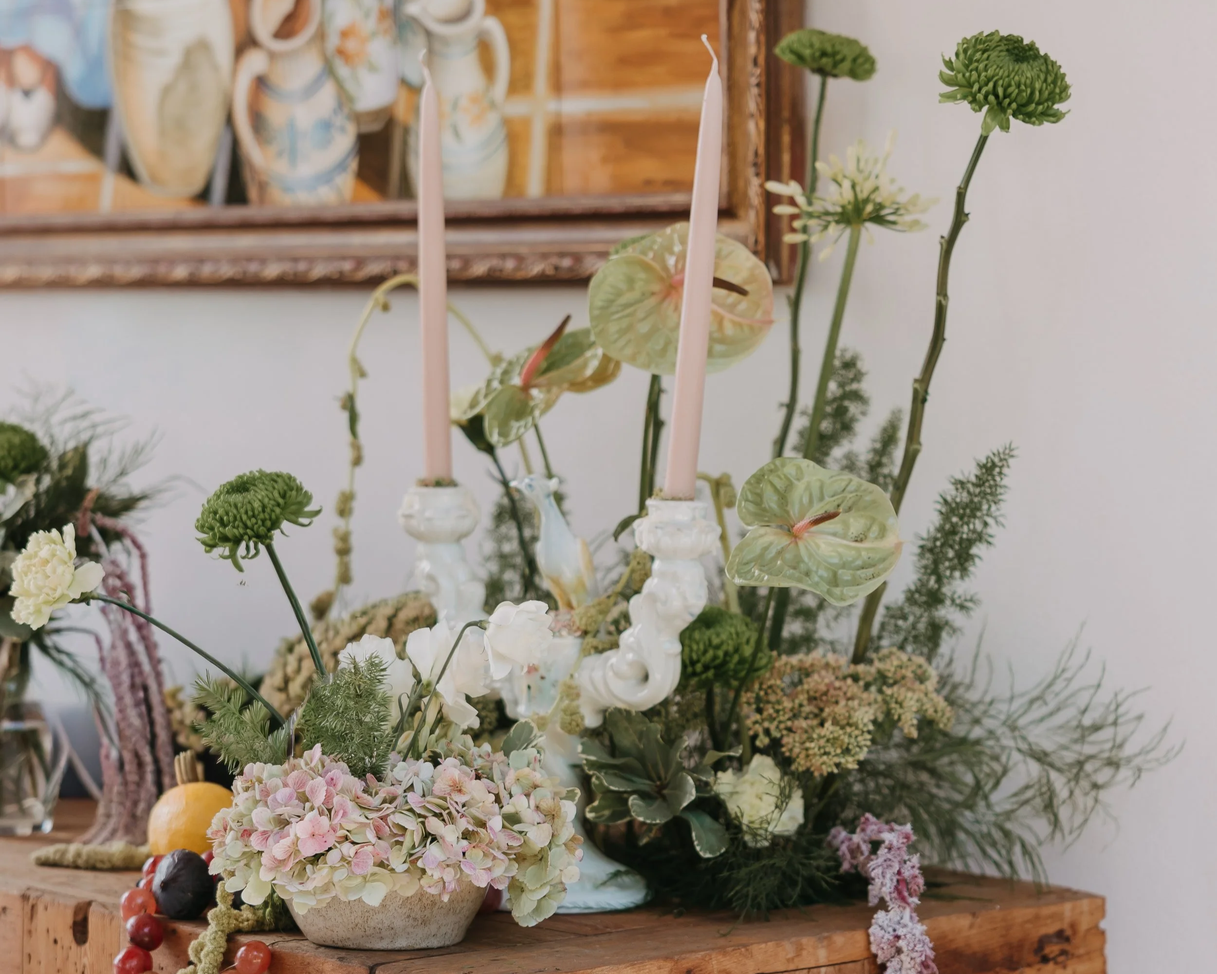 An arrangement of flowers and greenery with a white chandelier-style candle holder and pink candles on a wooden surface.