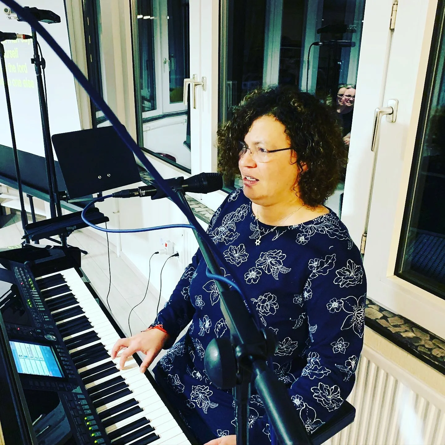 Eine Frau spielt Klavier und singt in einem Raum vor einem Mikrofon, im Hintergrund sind Fenster und eine Person, die zuschaut.