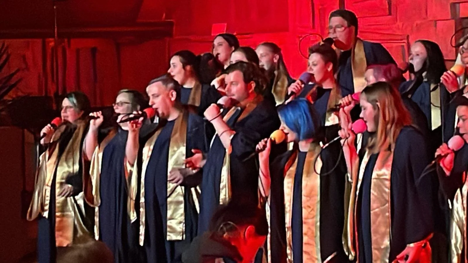 Chor mit mehreren singenden Mitgliedern, die Mikrofone halten, auf einer Bühne mit rotem Hintergrund, alle tragen schwarze Roben mit goldenen Applikationen.