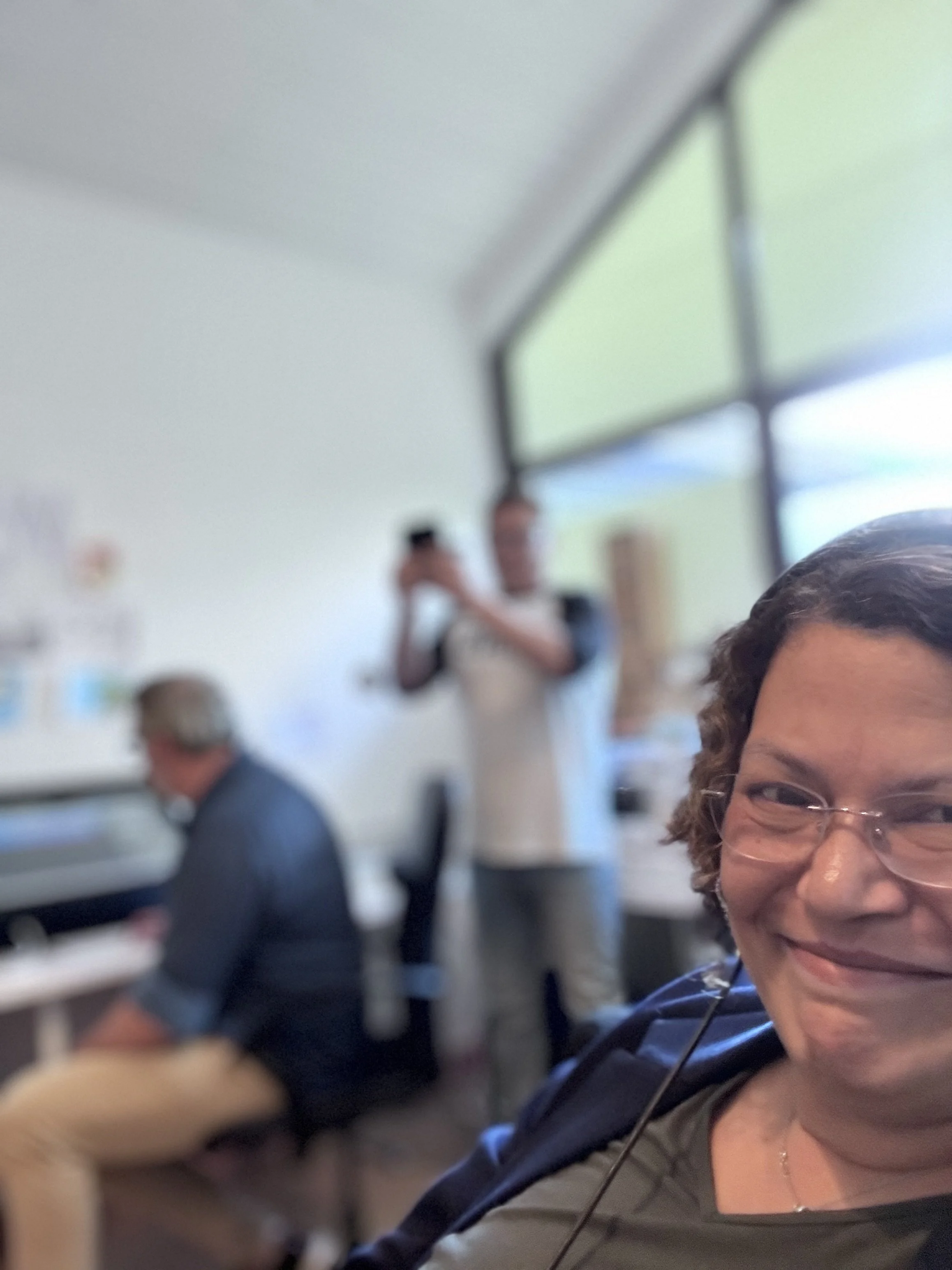 Eine Frau mit Brille lächelt in die Kamera. Im Hintergrund sind zwei Männer, einer sitzt an einem Tisch und der andere steht und fotografiert.