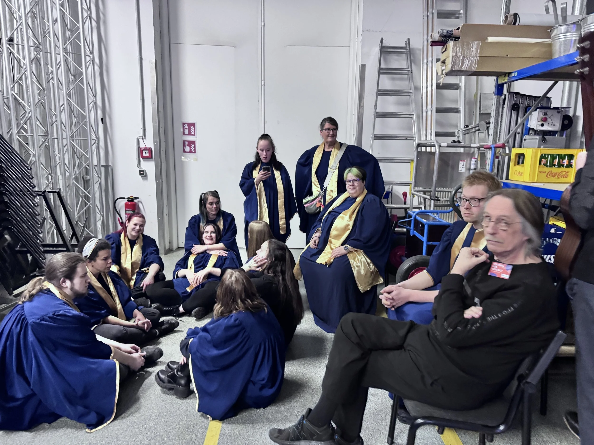 Gruppe von Menschen in blauen Roben mit goldenen Schals, some sitzen auf dem Boden, andere sitzen oder stehen im Raum, hinter ihnen Regale, Leiter und Getränkeablagen.