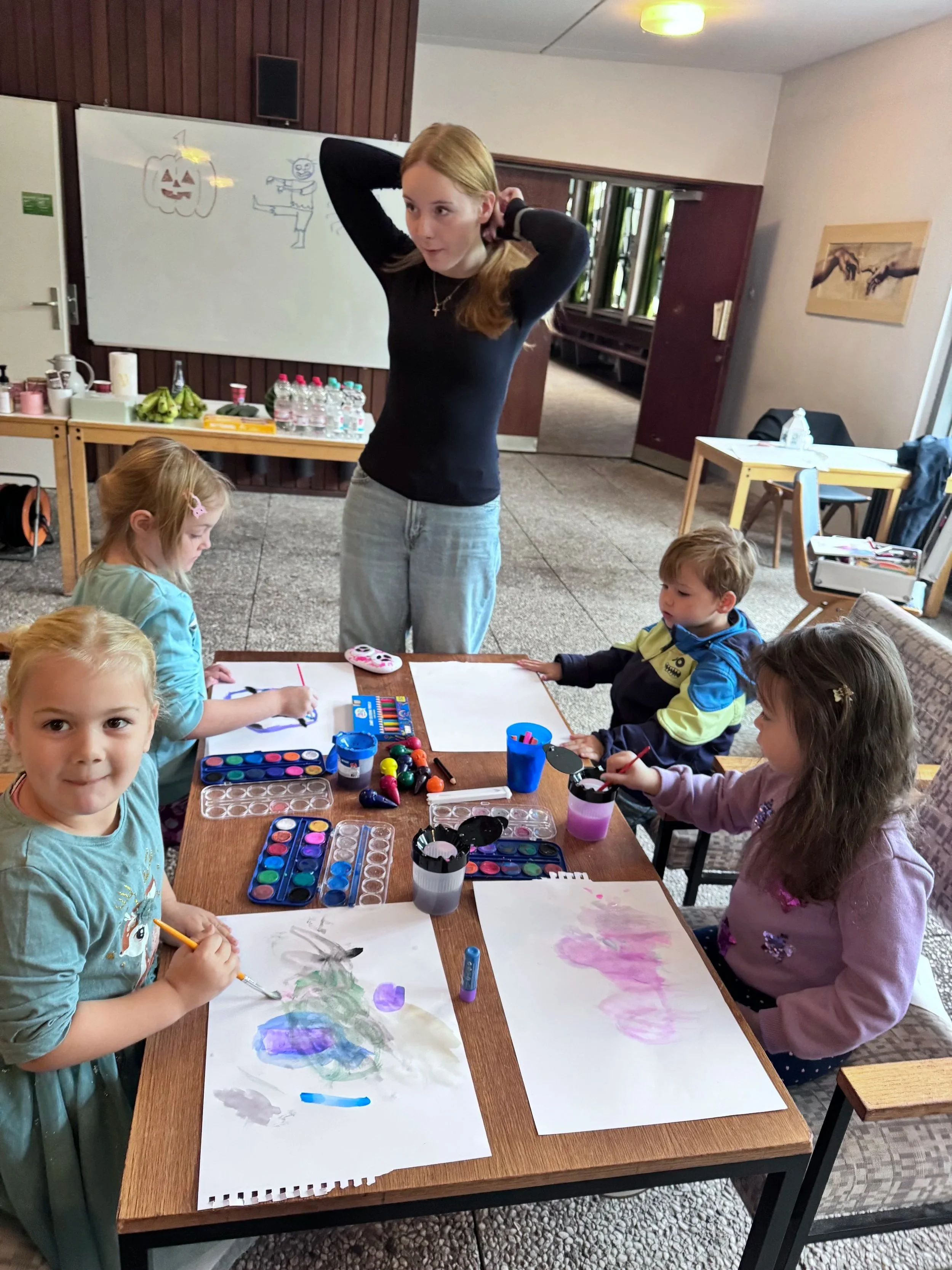 Eine Betreuerin unterrichtet vier Kinder beim Malen an einem Tisch in einem Raum.