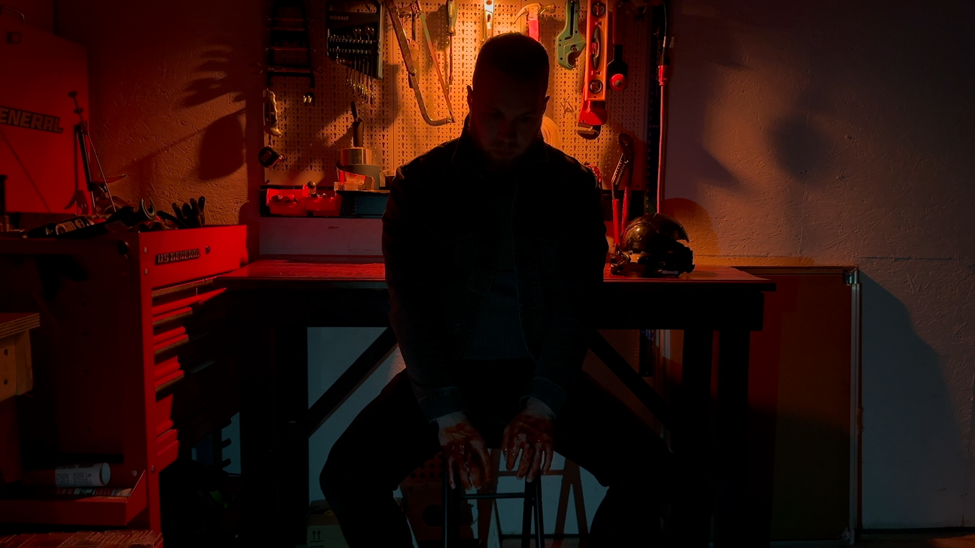 A person sitting at a workbench in a dimly lit workshop, with tools hanging on a pegboard behind them, and a helmet on the work surface. The person's face is not visible, and the scene is illuminated with warm, low lighting.