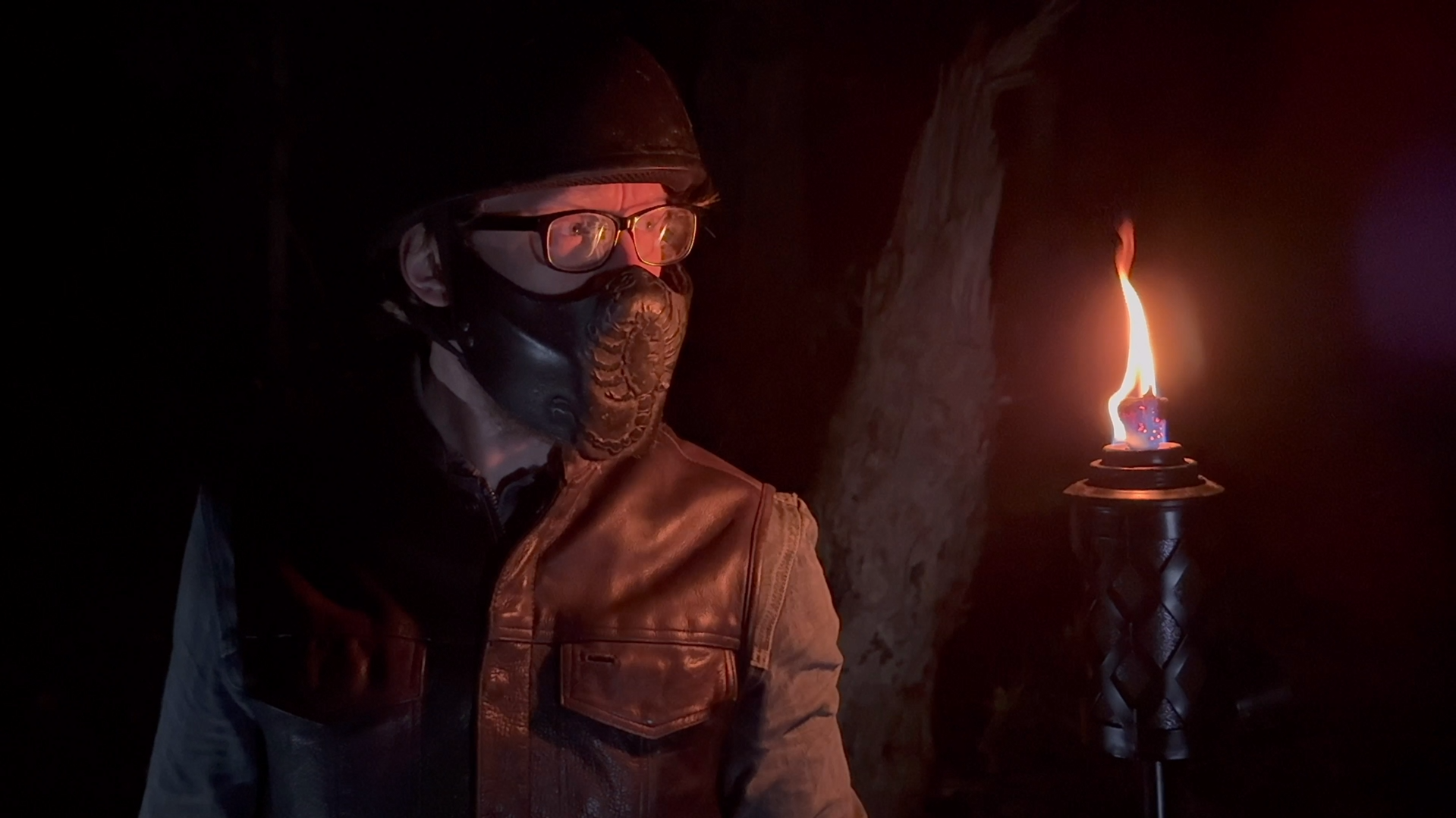 A person wearing glasses, a black face mask, a dark helmet, and a brown leather jacket standing in darkness illuminated by a nearby torch with a flame.