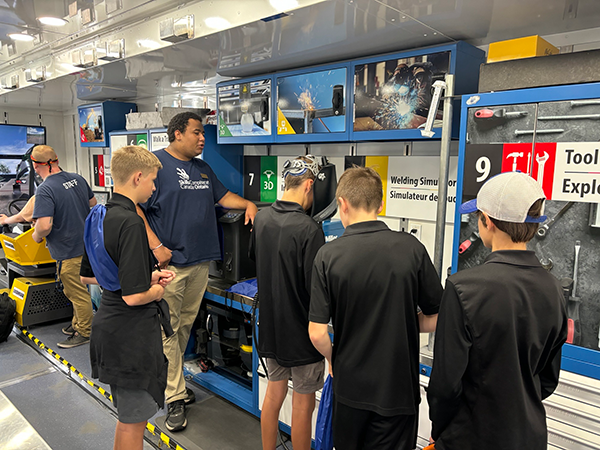Group of young people learning welding simulation at a TIG welding educational station with monitors and equipment in a workshop setting.