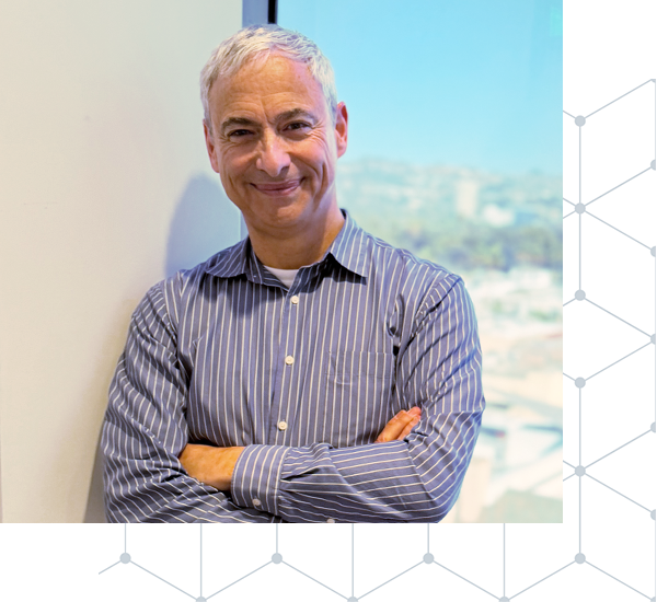 A smiling middle-aged man with gray hair, wearing a blue striped button-up shirt, standing with arms crossed beside a white wall with a window in the background showing a city landscape.